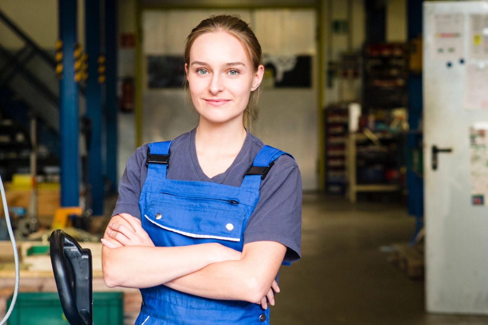 Eine junge Frau im Betrieb in einer blauen Arbeitshose und einem blauen T-Shirt steht mit verschränkten Armen da und schaut freundlich