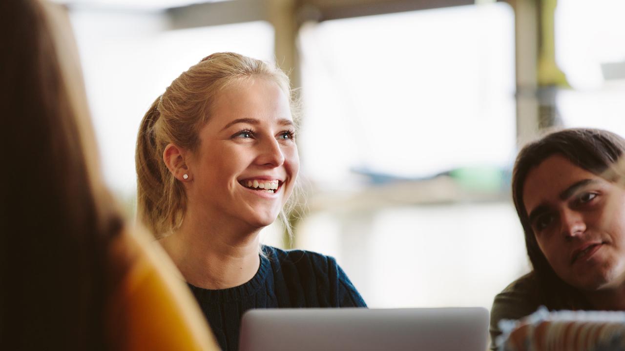 Eine Junge Frau sitzt vor einem Laptop und unterhält sich mit anderen jungen Leuten